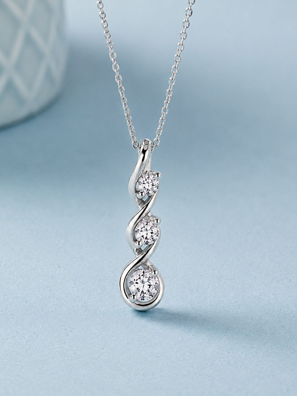 Journey moissanite pendant necklace hanging upright, 3 graduated round stones 3mm to 5mm in a twisted curve setting, 0.75 CTW, rhodium plated 925 sterling silver, on light blue background – Adora Fine Jewelry