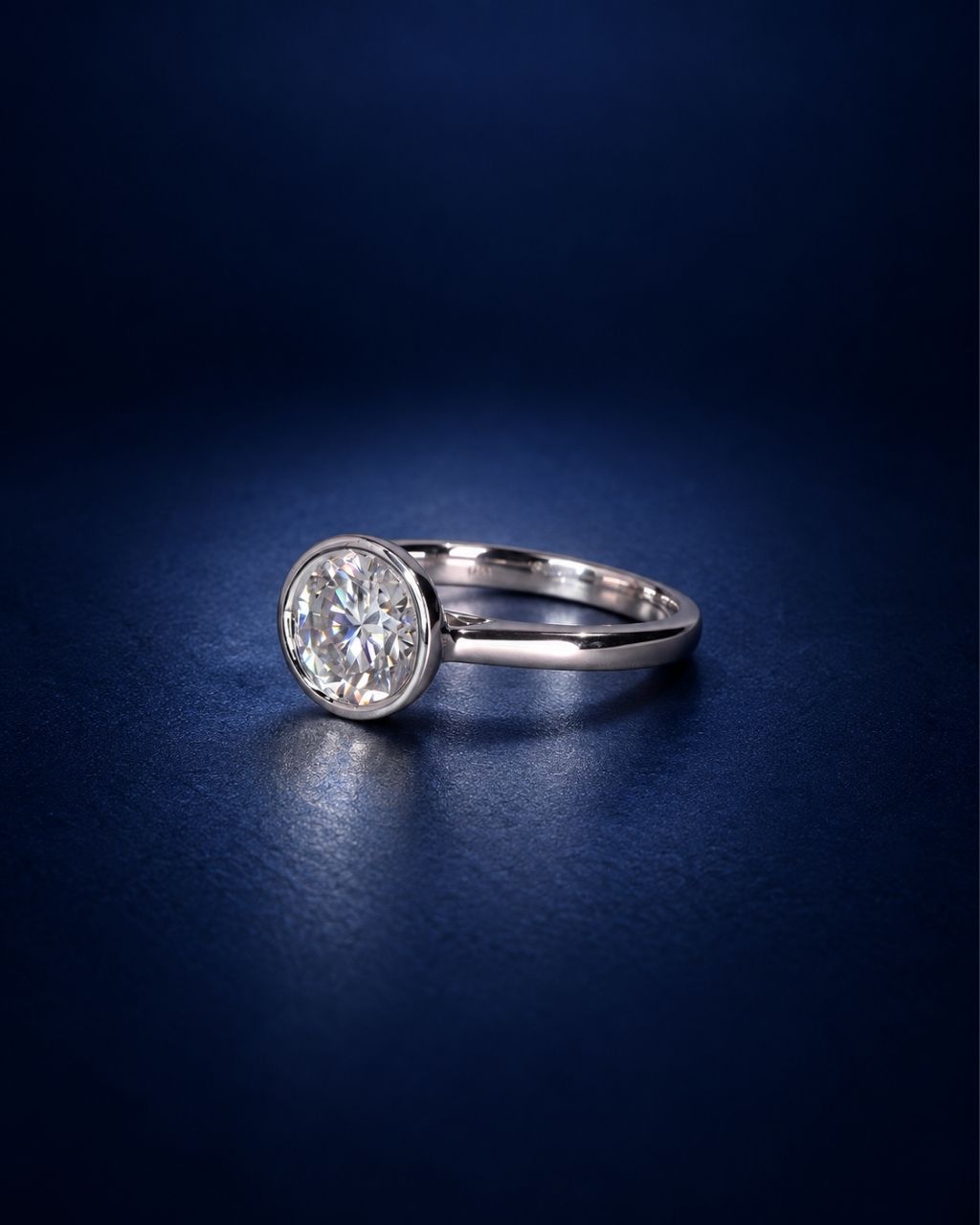 Round bezel-set moissanite solitaire ring in polished 14K solid white gold, shown at a 45-degree angle on a deep navy blue background