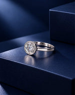 Round bezel-set moissanite solitaire ring in polished 14K solid white gold, elegantly displayed on a textured navy blue platform with dramatic spotlighting