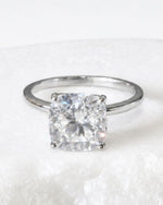 A cushion-cut diamond ring in a silver four-prong setting, resting on a textured white surface.