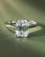 An emerald-cut diamond ring in a silver four-prong setting, resting on a textured olive green surface.