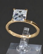 A yellow gold solitaire ring featuring a princess-cut clear gemstone, displayed on a clear acrylic stand against a dark gray background.