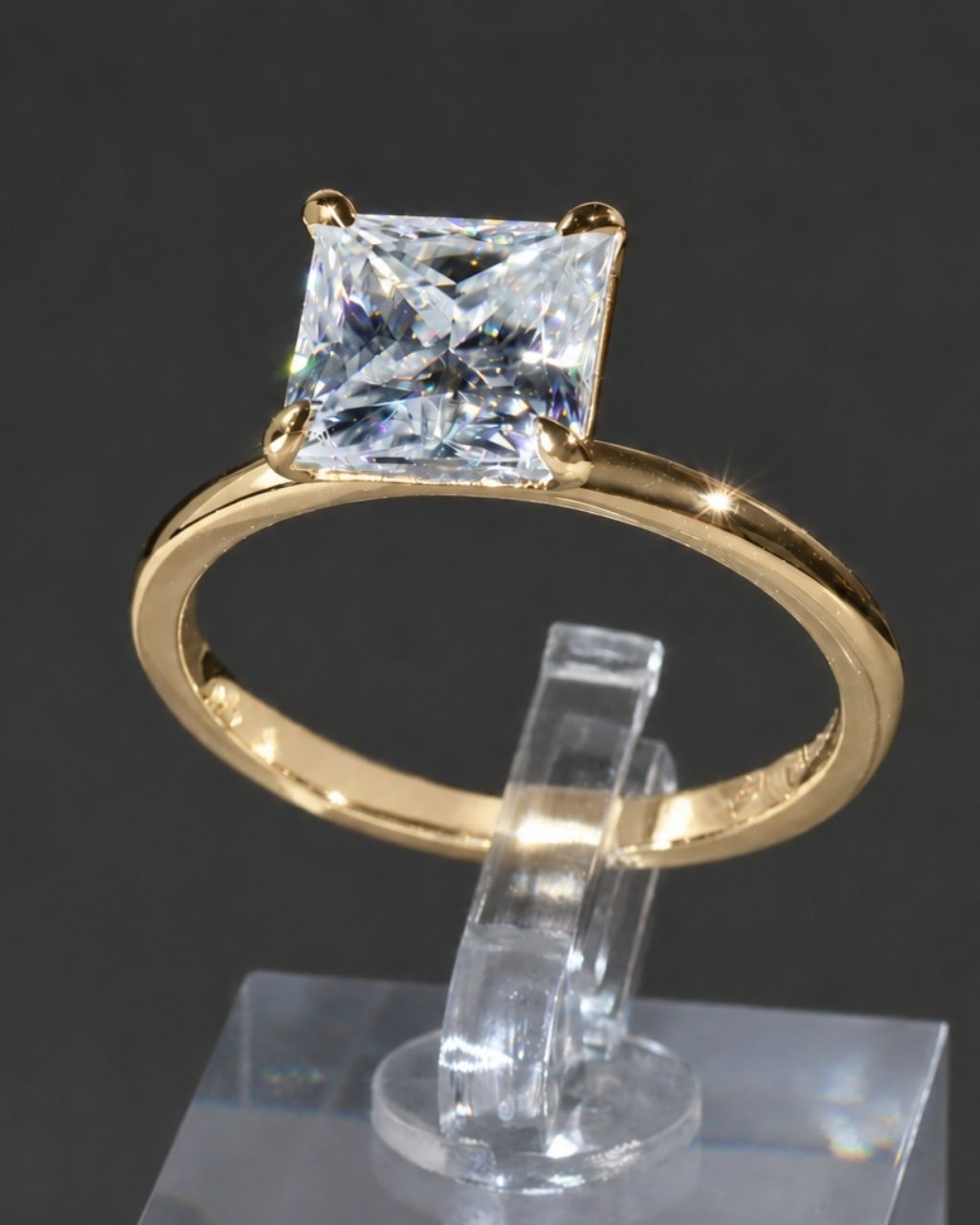 A yellow gold solitaire ring featuring a princess-cut clear gemstone, displayed on a clear acrylic stand against a dark gray background.