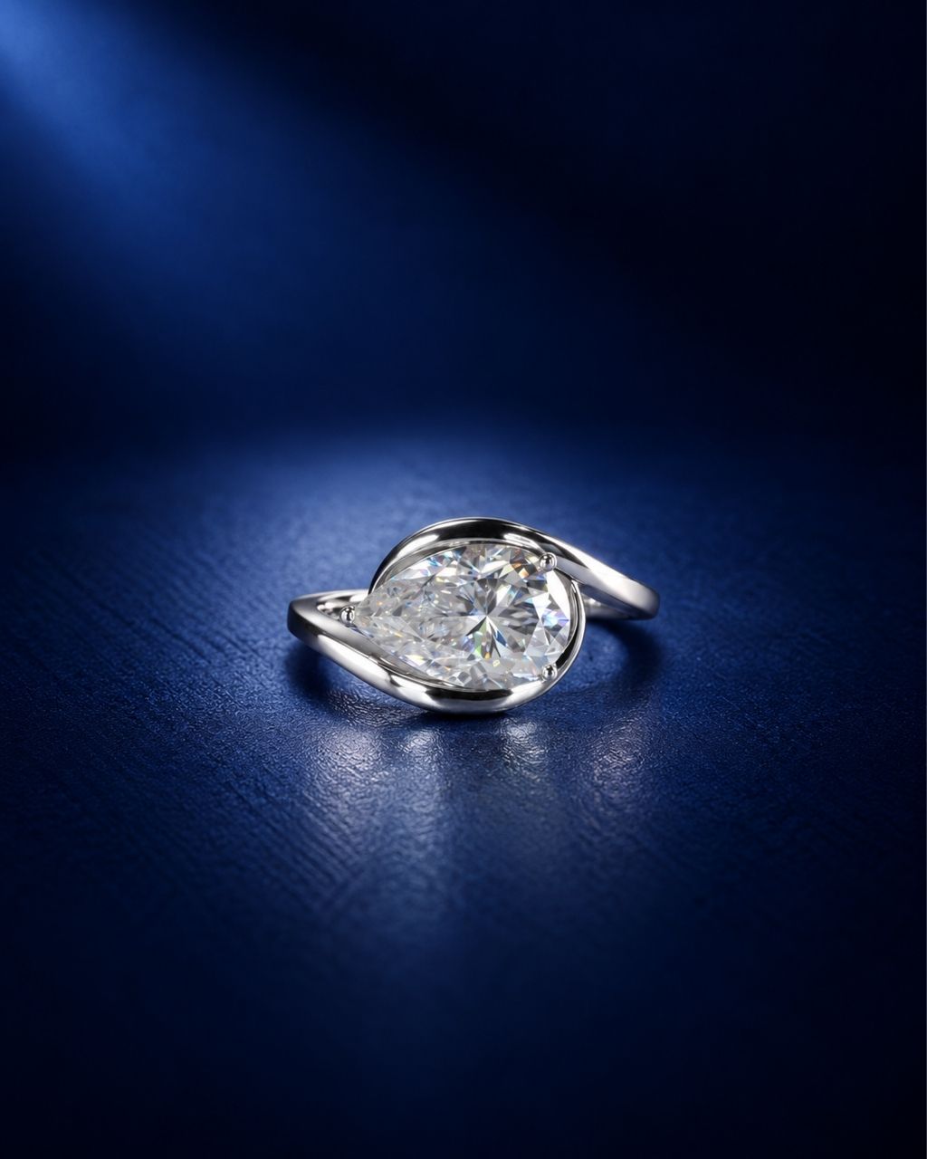 Pear-cut moissanite engagement ring with a polished white gold swirl bypass setting, photographed on a deep navy blue background.
