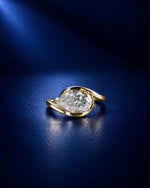 Pear-cut moissanite engagement ring in polished yellow gold with an elegant swirl bypass setting, photographed on a deep navy blue background