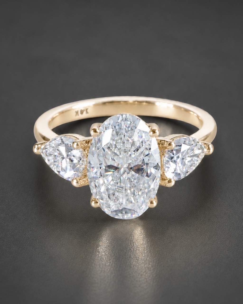 A yellow gold three-stone ring featuring a large oval center diamond flanked by two smaller pear-cut diamonds, photographed on a dark gray reflective surface.