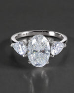 A silver three-stone diamond ring featuring a large oval center diamond flanked by two smaller pear-cut diamonds, set against a dark gray reflective background.