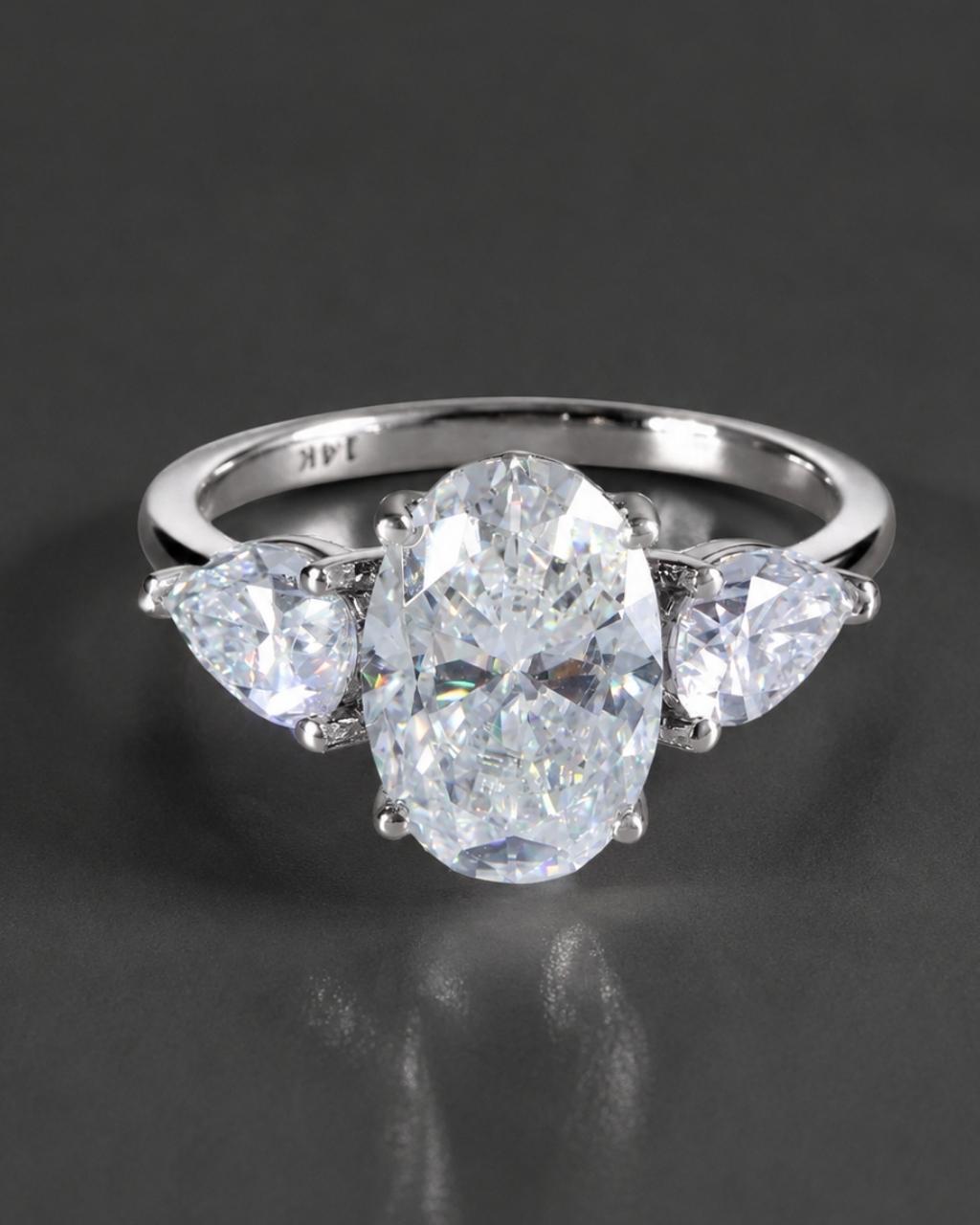 A silver three-stone diamond ring featuring a large oval center diamond flanked by two smaller pear-cut diamonds, set against a dark gray reflective background.